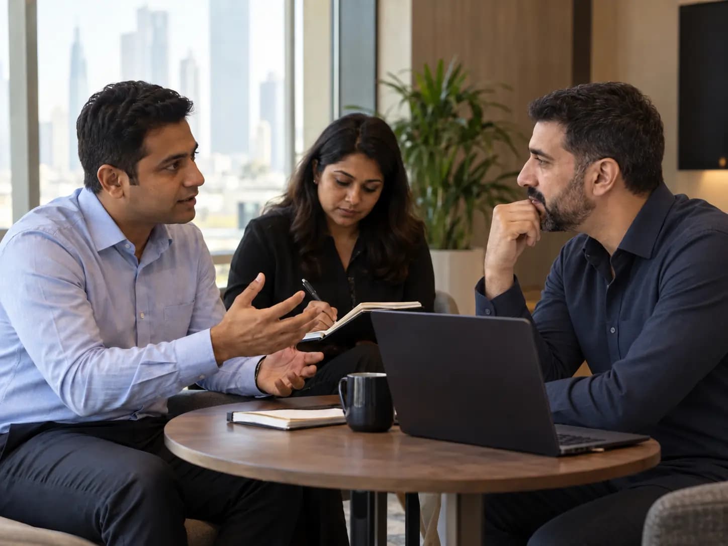 Three colleagues talking around a table, one speaking while the others listen and take notes.